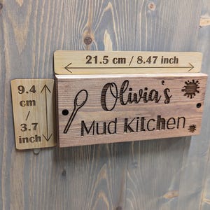 May include: A wooden sign reading "Olivia's Mud Kitchen" with a spoon illustration. The sign measures 21.5 cm / 8.47 inches wide and 9.4 cm / 3.7 inches high. The sign is made of light brown wood and is mounted on a grey wooden surface.