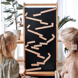 Op de afbeelding: Een houten educatief speelgoed met een zwart oppervlak en lichtgekleurde houten blokken in een zigzagpatroon. Het frame is gemaakt van lichtgekleurd hout. Twee jonge kinderen kijken naar het speelgoed.