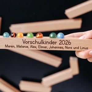 Könnte beinhalten: Eine Holzkugelbahn mit bunten Murmeln und dem Text "Vorschulkinder 2026" und Namen. Die Murmeln sind blau, orange, grün und rot. Die Holzstrecke wird von einer Hand gehalten.