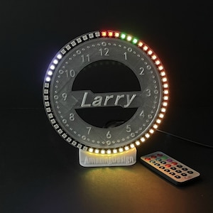 Puede incluir: Una luz LED circular con una base blanca y un control remoto. La luz tiene una esfera de reloj con los números del 1 al 12 y el nombre "Larry" impreso en el centro. La luz se ilumina en una variedad de colores, incluyendo rojo, verde, amarillo y blanco.