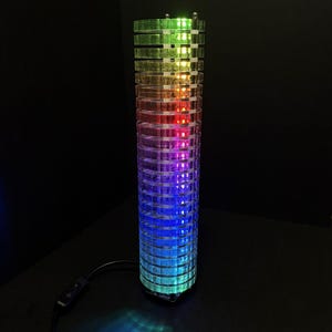 Op de afbeelding: Een hoge, cilindrische lamp met een regenboogkleurenverloop. De lamp is gemaakt van heldere, gesegmenteerde stukken waardoor de interne LED-verlichting doorschijnt. De kleuren gaan over van groen naar blauw, met rood en paars in het midden.