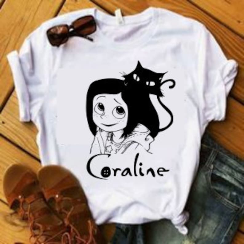 May include: White t-shirt with a black and white illustration of Coraline from the movie Coraline, with the black cat from the movie behind her. The text "Coraline" is printed in black below the illustration.