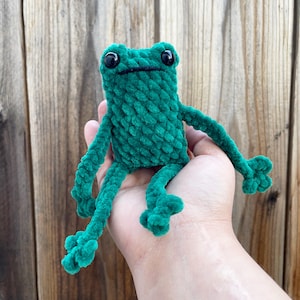 May include: A green crocheted frog plush toy with black eyes. The frog is sitting on a person's hand.