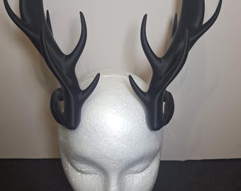 May include: Black antler headband with a detailed, realistic design. The antlers are mounted on a black headband base. The headpiece is displayed on a white foam head, showcasing the shape and style of the accessory.
