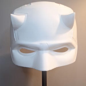 May include: A white superhero mask with distinctive horns and eye openings. The mask is mounted on a black stand against a neutral backdrop. The design features a defined brow and a textured surface.