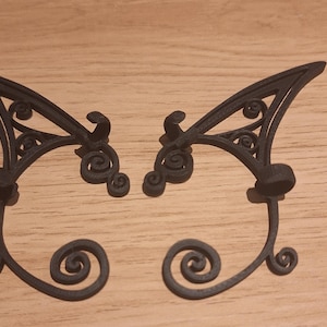 May include: Pair of black ear cuffs with an intricate, filigree design. The ear cuffs have a curved shape with a swirling, scroll-like pattern at the bottom and top. The ear cuffs are made of a dark material and are designed to be worn on the ear.