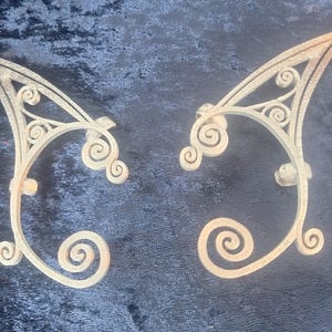 May include: A pair of light-coloured metal ear cuffs with an ornate, fantasy design. Each cuff has a curved shape with intricate scrollwork and a pointed top, designed to clip onto the ear. The cuffs are displayed on a dark blue velvet surface.