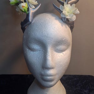 May include: A silver-colored antler headband adorned with white and green artificial flowers. The headpiece is displayed on a white mannequin head, showcasing the design and floral accents. The headband is a unique accessory.