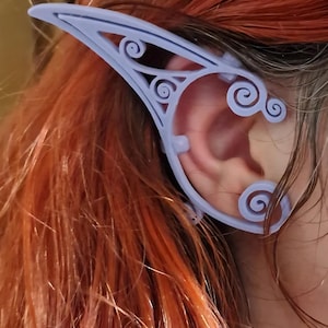 May include: A pale blue elf ear prosthetic, 3D-printed with a decorative, swirling pattern. The prosthetic is worn over the ear, with red hair visible in the background.
