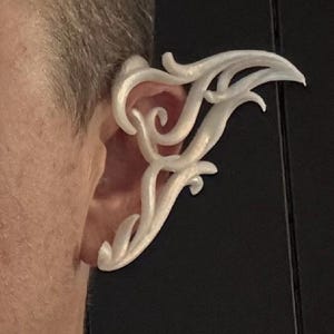 May include: A white, ornate ear cuff with a flowing, stylized design. The ear accessory curves around the ear, featuring swirling and pointed elements. The cuff is worn on a person's ear, with the skin visible.