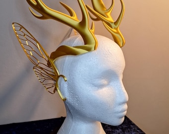 May include: A golden headpiece featuring deer antlers and delicate fairy wings. The headpiece is displayed on a white mannequin head, showcasing the intricate details and design. The antlers are approximately 6 inches tall.
