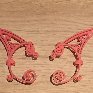 May include: A pair of coral-coloured elf ear cuffs. Each cuff has an intricate, openwork design with swirling patterns and a curved shape. The cuffs are designed to clip onto the ear, adding a whimsical touch to any look.