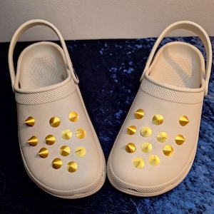 Könnte beinhalten: Ein Paar beigefarbene Clogs mit goldfarbenen, kegelförmigen Nieten. Die Clogs haben einen Riemen, der um die Ferse oder über dem Fuß getragen werden kann. Die Schuhe sind aus einem gummiartigen Material gefertigt und für den Freizeitgebrauch konzipiert.