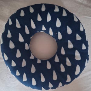 May include: A navy blue donut-shaped pillow featuring a white raindrop design. The pillow has a circular opening in the centre. The pillow is made of a soft fabric.