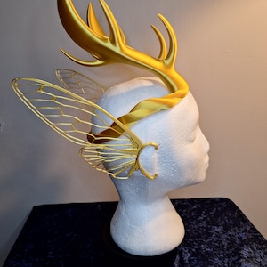 May include: A gold-coloured headpiece with antler-like structures and insect wing details. The headpiece is displayed on a white mannequin head, highlighting its intricate design. The headpiece is made of a shiny gold material.