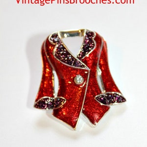 May include: A red enamel brooch shaped like a jacket with silver trim and purple rhinestone accents. The brooch has a single clear rhinestone on the front.