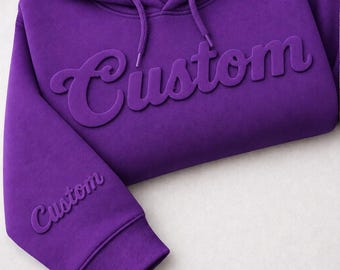 May include: A vibrant purple hooded sweatshirt featuring the word Custom in a raised, embossed script font across the chest and a smaller version of the same embossed text on the lower right sleeve, complete with a matching drawstring hood.