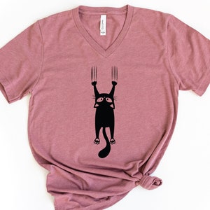 May include: A mauve v-neck t-shirt with a black silhouette of a cat hanging upside down with its claws extended.