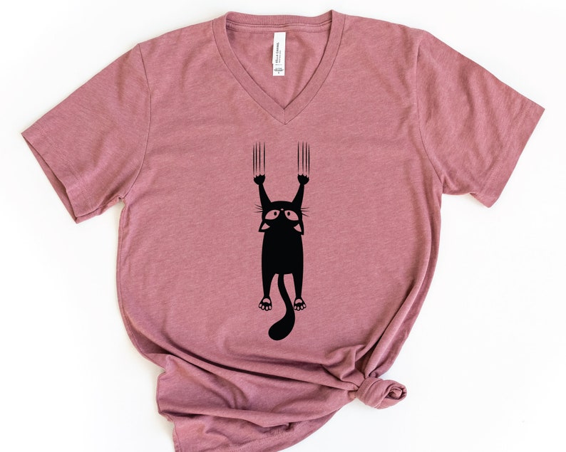 May include: A mauve v-neck t-shirt with a black silhouette of a cat hanging upside down with its claws extended.
