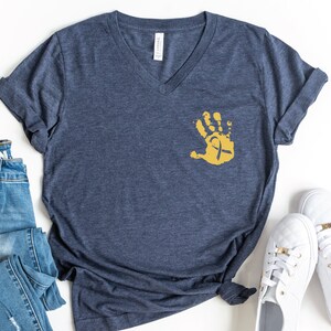 May include: A navy blue v-neck t-shirt with a gold handprint and ribbon design. The shirt is laid flat on a white surface with a pair of white sneakers and a pair of blue jeans.