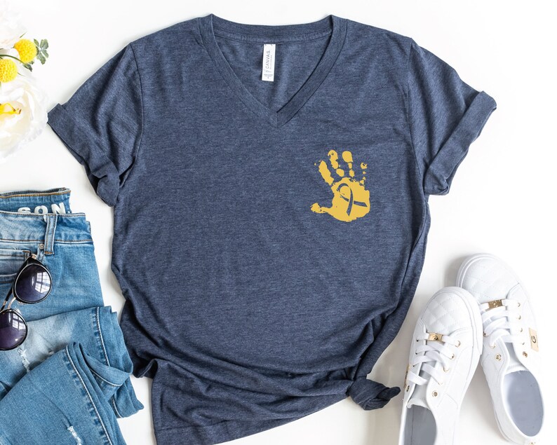 May include: A navy blue v-neck t-shirt with a gold handprint and ribbon design. The shirt is laid flat on a white surface with a pair of white sneakers and a pair of blue jeans.