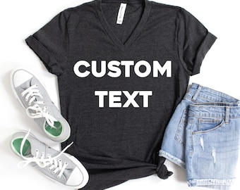 May include: A dark heather gray V-neck t-shirt with short sleeves and the white text CUSTOM TEXT printed on the front, accompanied by a pair of light blue denim shorts with frayed hems and two light gray low-top canvas sneakers with white laces.