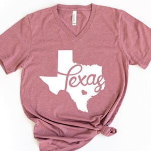 Puede incluir: Una camiseta de cuello en V de color malva con un contorno blanco del estado de Texas y la palabra "Texas" en una fuente blanca cursiva.