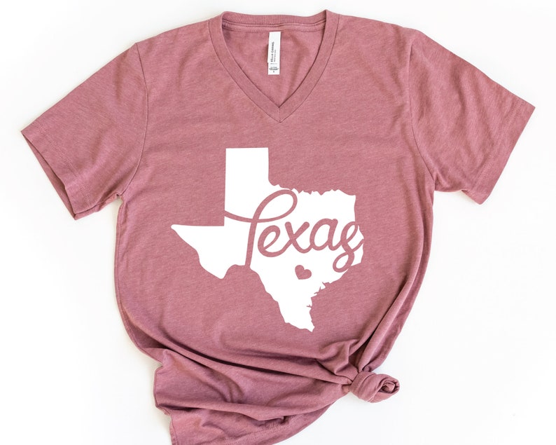 Puede incluir: Una camiseta de cuello en V de color malva con un contorno blanco del estado de Texas y la palabra "Texas" en una fuente blanca cursiva.