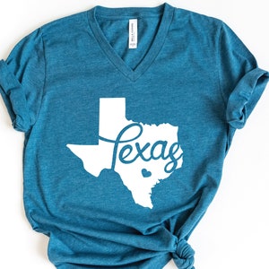 Puede incluir: Una camiseta azul turquesa con un contorno blanco del estado de Texas y la palabra "Texas" escrita en letra cursiva dentro del contorno.