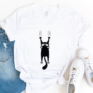 May include: White v-neck t-shirt with a black silhouette of a cat hanging from a surface with its claws extended.