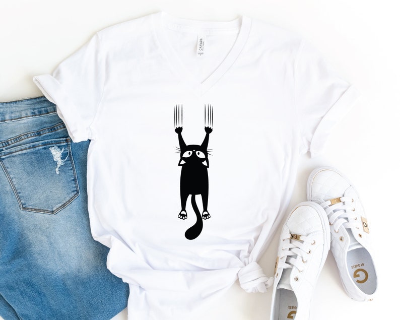 May include: White v-neck t-shirt with a black silhouette of a cat hanging from a surface with its claws extended.