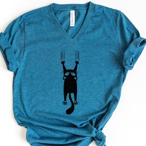 May include: A teal blue v-neck t-shirt with a black graphic of a cat hanging upside down with its claws extended.