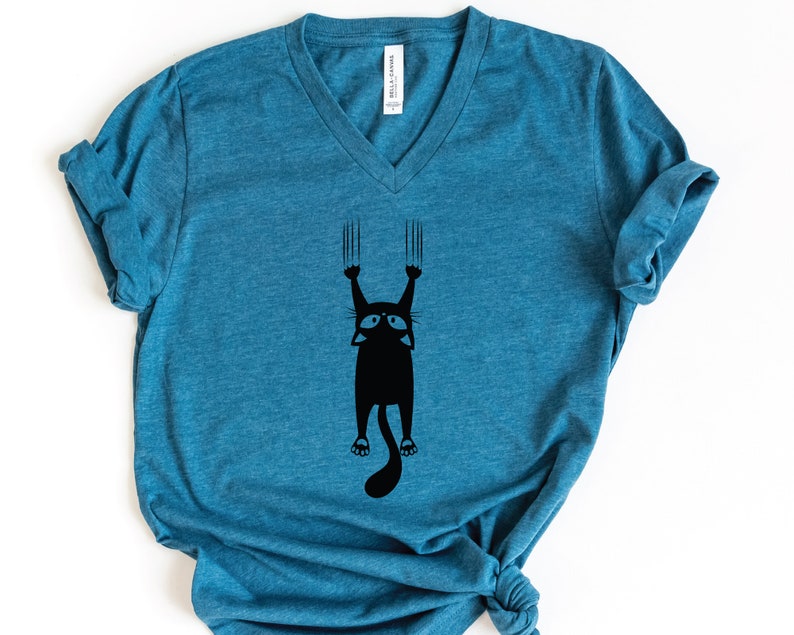 May include: A teal blue v-neck t-shirt with a black graphic of a cat hanging upside down with its claws extended.