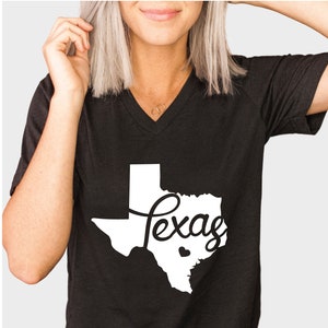 Puede incluir: Camiseta negra de cuello en V con un contorno blanco del estado de Texas y la palabra "Texas" escrita en letra cursiva dentro del contorno. Un peque&ntilde;o coraz&oacute;n blanco est&aacute; dentro del contorno del estado.