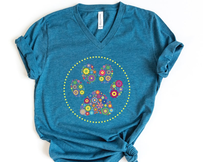 May include: A teal blue t-shirt featuring a floral paw print design. The paw print is made up of colourful flowers and is surrounded by a yellow dotted circle.