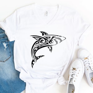 May include: White v-neck t-shirt with a black tribal shark design. The shirt is laid flat on a white surface with a pair of white sneakers next to it.