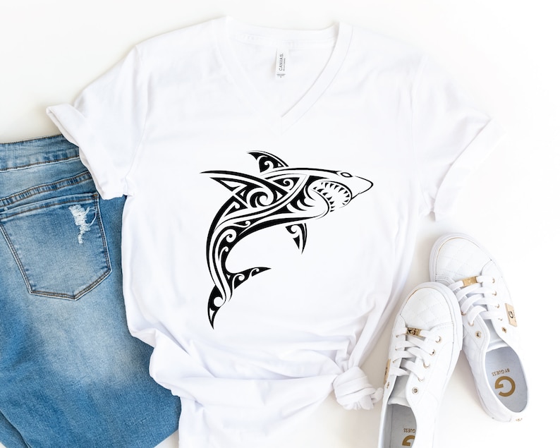 May include: White v-neck t-shirt with a black tribal shark design. The shirt is laid flat on a white surface with a pair of white sneakers next to it.