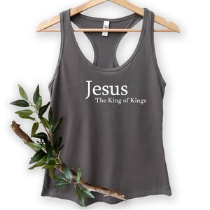 May include: Gray racerback tank top with the words "Jesus The King of Kings" printed in white. The tank top is hanging on a wooden hanger. A branch with green leaves is placed in front of the tank top.