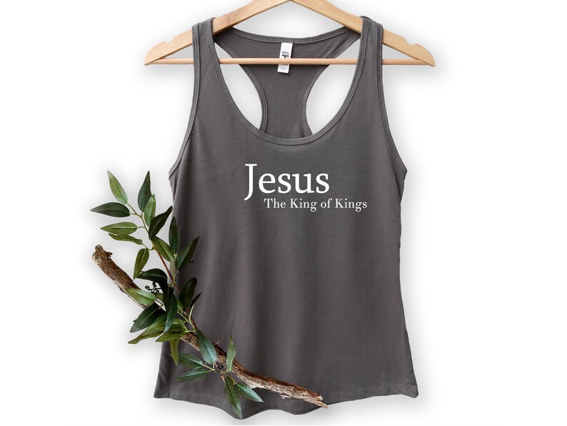 May include: Gray racerback tank top with the words "Jesus The King of Kings" printed in white. The tank top is hanging on a wooden hanger. A branch with green leaves is placed in front of the tank top.