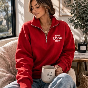May include: A red sweatshirt with a quarter-zip neckline and the text "YOUR LOGO HERE" in white. The sweatshirt is paired with blue jeans. A ceramic mug with the words "good days" is held in the hand.