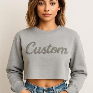 A young woman with brown hair wearing a gray sweatshirt with the word "Custom" printed on it, standing against a plain background.