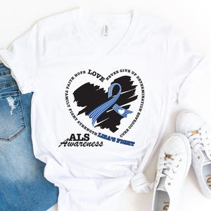 May include: A white V-neck t-shirt featuring a black heart graphic with a blue striped awareness ribbon. The design includes the text: FAITH HOPE LOVE NEVER GIVE UP DETERMINATION FAMILY FIGHT STRENGTH CURE COURAGE, ALS AWARENESS, and LISA'S FIGHT.