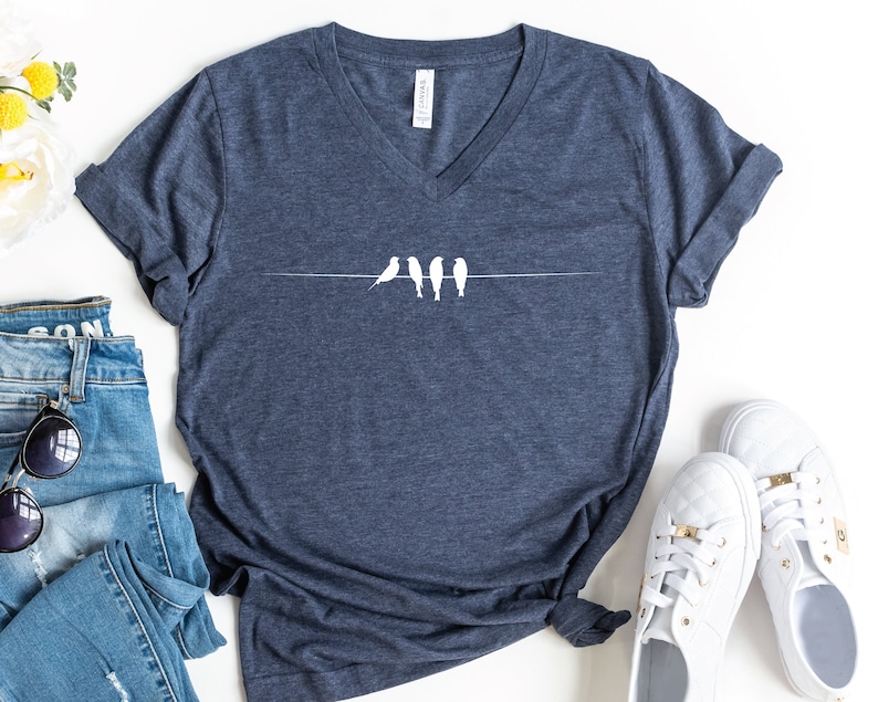Pu&ograve; includere: Una maglietta blu navy a collo a V con una grafica bianca di cinque uccelli appollaiati su un filo. La maglietta &egrave; stesa su una superficie bianca con un paio di scarpe da ginnastica bianche e un paio di jeans blu.
