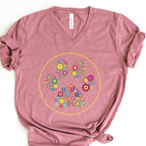 May include: A mauve v-neck t-shirt featuring a floral paw print design. The paw print is made up of colourful flowers and is surrounded by a circle of yellow dots.