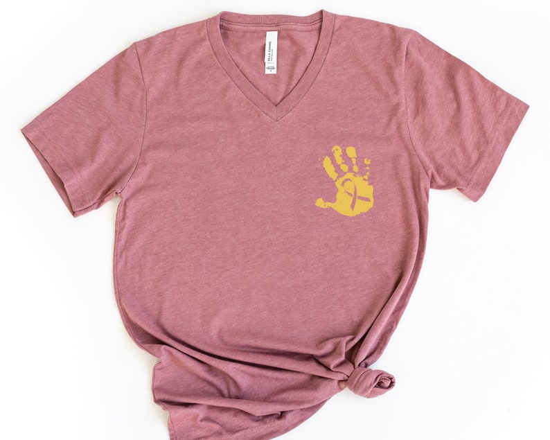 May include: A mauve v-neck t-shirt with a gold handprint design featuring a yellow ribbon. The handprint is on the left side of the shirt.