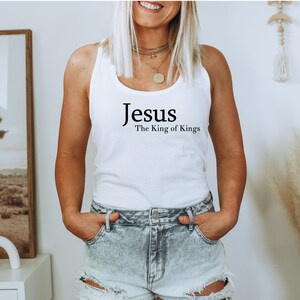 May include: White tank top with the text "Jesus The King of Kings" in black. The top is a simple, sleeveless design, perfect for casual wear. The image shows the top on a person.
