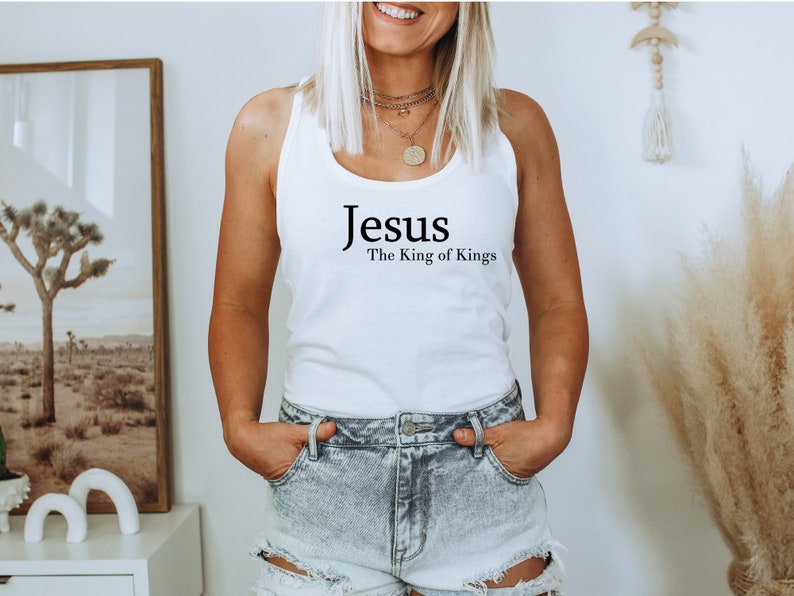 May include: White tank top with the text "Jesus The King of Kings" in black. The top is a simple, sleeveless design, perfect for casual wear. The image shows the top on a person.