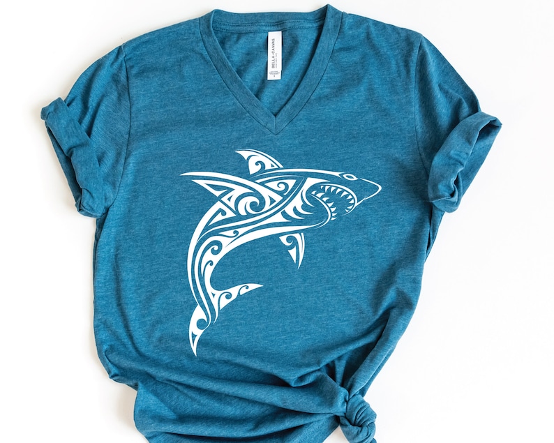 May include: A teal blue t-shirt with a white tribal shark design. The shark is facing right and has a detailed design with swirls and lines.