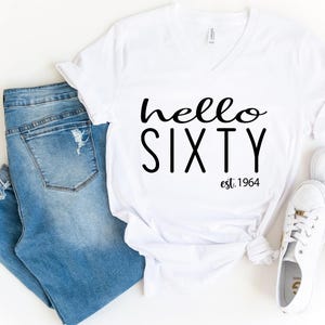 May include: A white V-neck t-shirt with the black text hello SIXTY est. 1964 printed on the front, paired with blue denim jeans featuring distressed details and a pair of white low-top sneakers with gold-toned eyelets and laces.