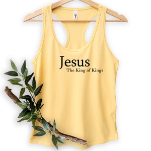 May include: A light yellow tank top with a racerback design. The text "Jesus The King of Kings" is printed in black on the front. The tank top is hanging on a wooden hanger. A branch with green leaves is placed in front of the tank top.